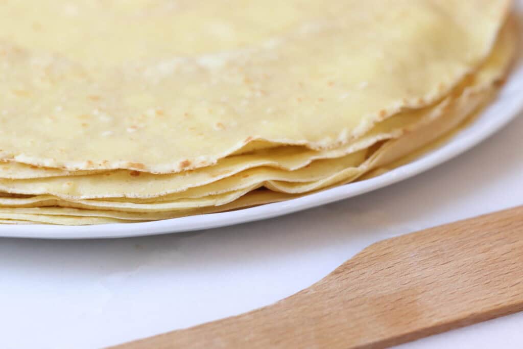 Thin French crepes stacked on a white plate with a wooden spatula nearby, showcasing classic French gouter or dessert options.