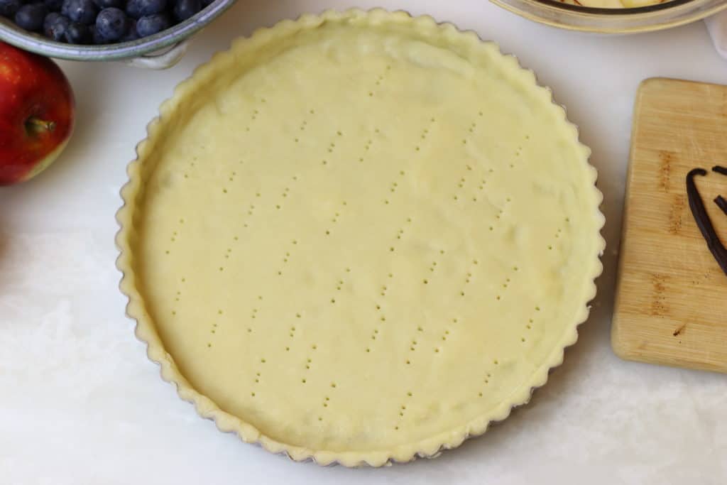 Unbaked Pâte Sucrée tart shell in tart pan, surrounding by apples and blueberries