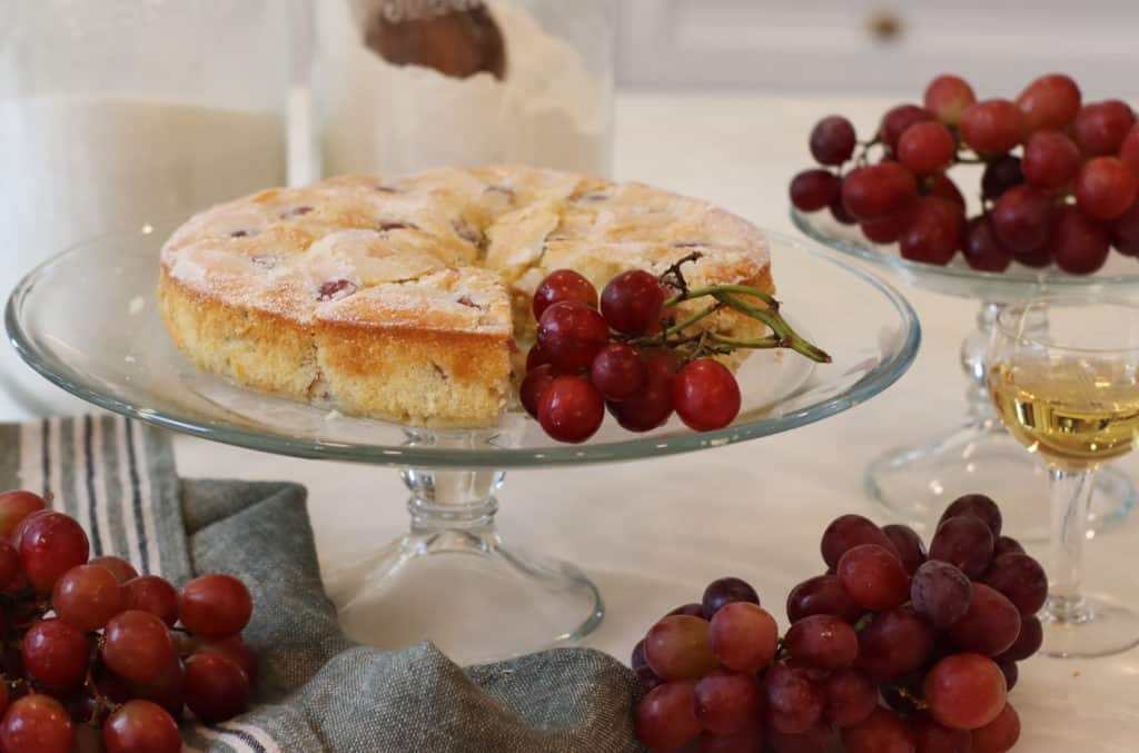 Grape cake on clear cake stand with clusters of grapes around it.