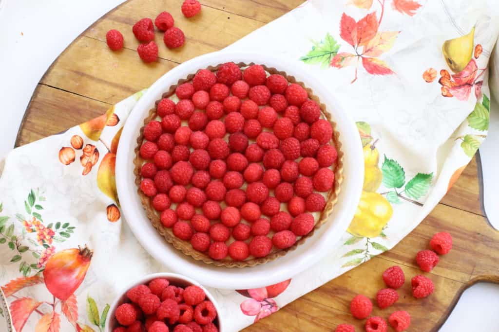 Raspberry tart on a tablecloth on top of cutting board.