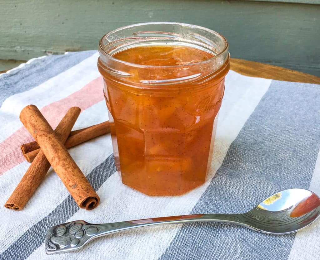 Open jar of jam sitting on a striped tablecloth with a spoon and cinnamon sticks near it.