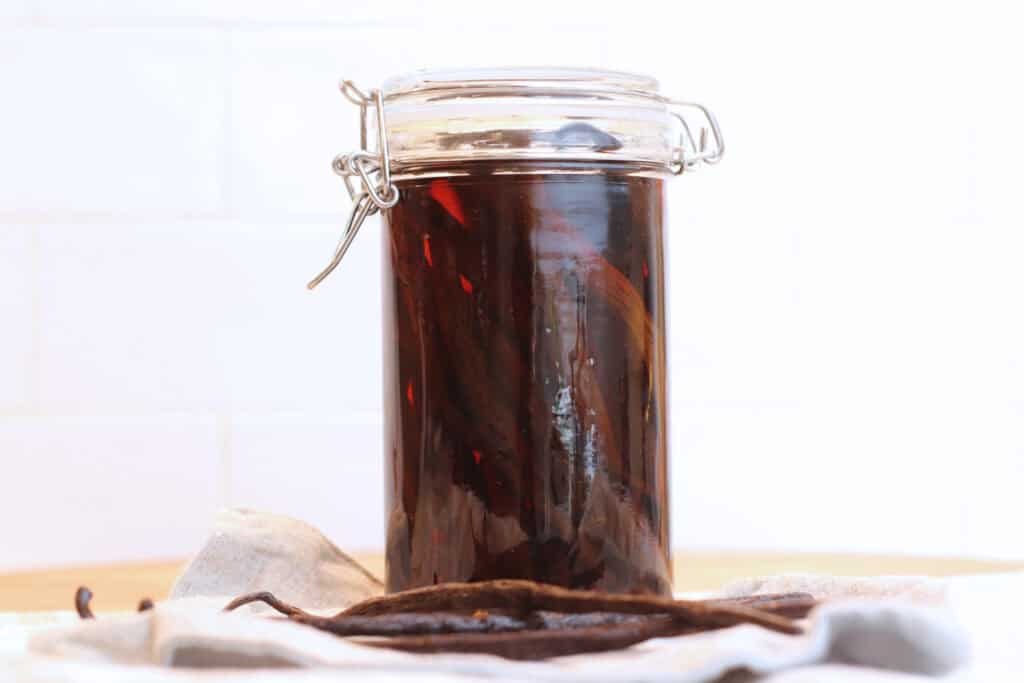 A clear jar filled with vanilla beans and homemade vanilla extract with loose vanilla beans in front against a white background.