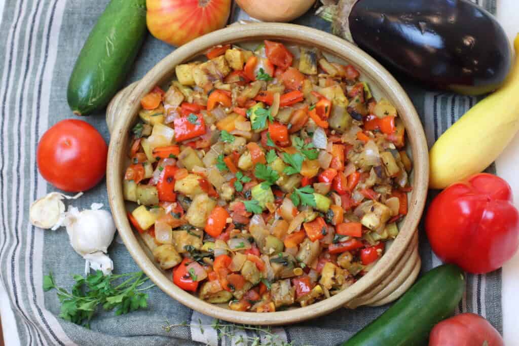 Ratatouille in a rustic brown serving dish on a tea towel surrounded by fresh vegetables.