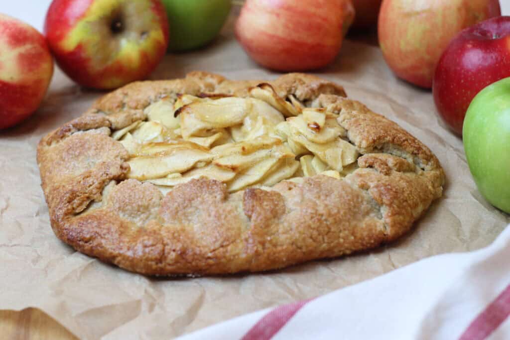 Freshly baked apple galette with a rustic crust, surrounded by colorful apples on a parchment paper-lined surface. Perfect for a cozy French-inspired dessert or pastry.