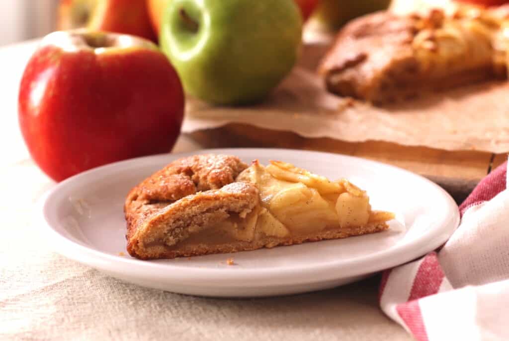 Fresh apple pie slice with baked apples on a white plate, surrounded by whole apples and homemade apple pie, perfect for a cozy French-inspired dessert or bakery presentation.