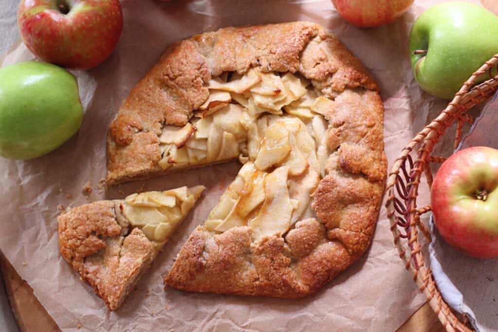 Golden apple galette with fresh apples on a rustic parchment paper background, surrounded by whole green and red apples, showcasing a classic French pastry perfect for fall desserts.