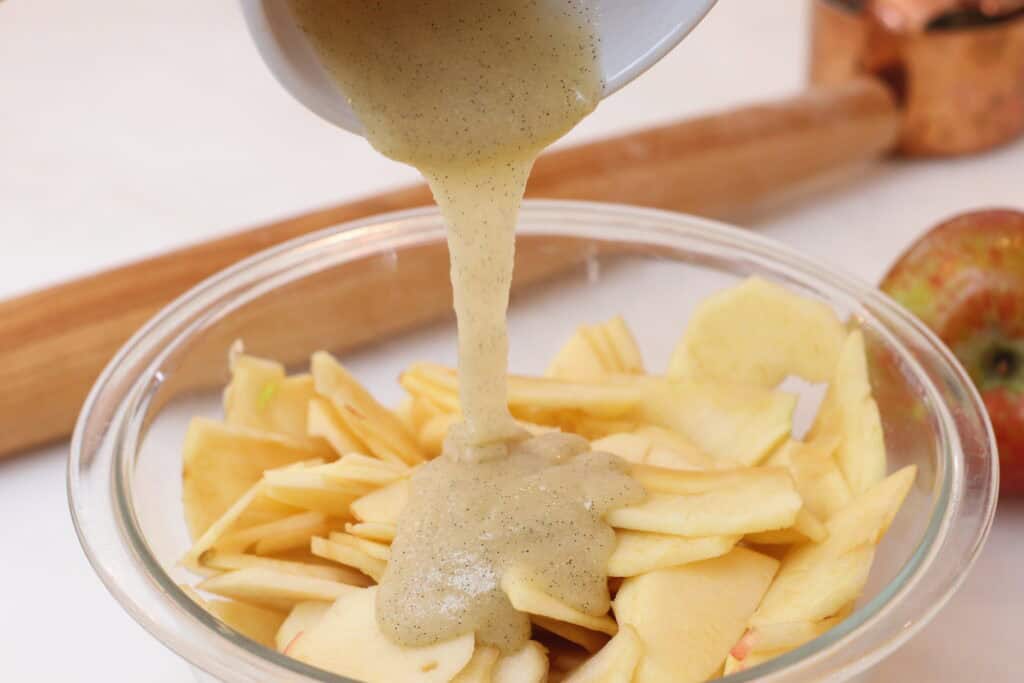 Vanilla lemon and sugar mixture being poured over sliced apples in a glass bowl, embodying classic French dessert traditions.