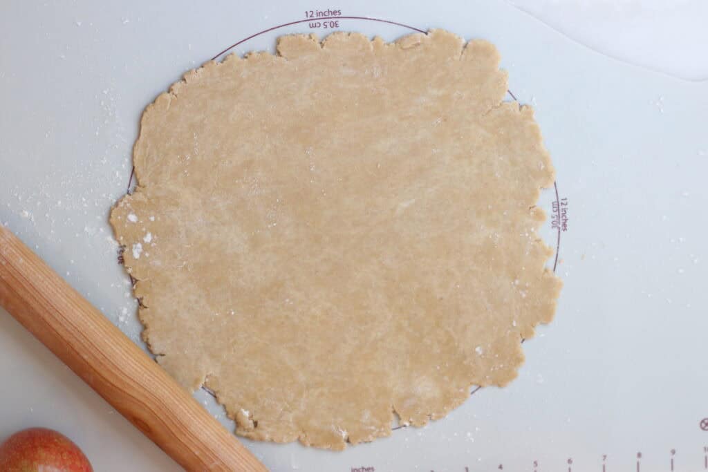 Fresh galette dough rolled out on a baking sheet with measurements next to a rolling pin and an apple.