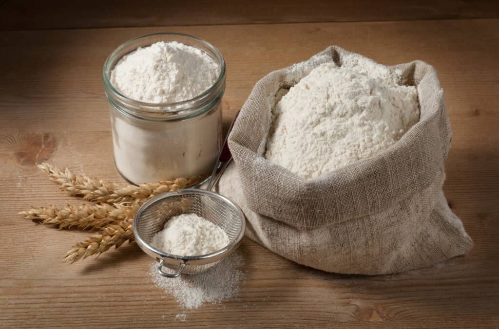 A sack of flour next to a jar of flour and sifter in a rustic French kitchen with wheat lying next to it.