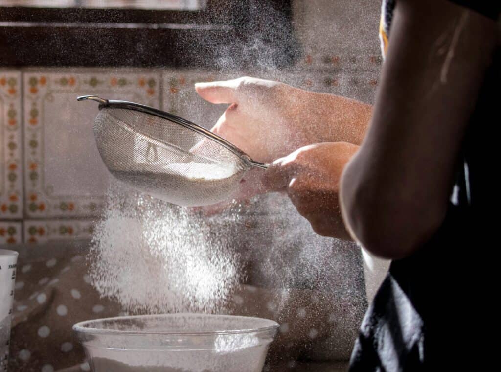 Hand sifting flour over a bowl in a rustic French kitchen setting.