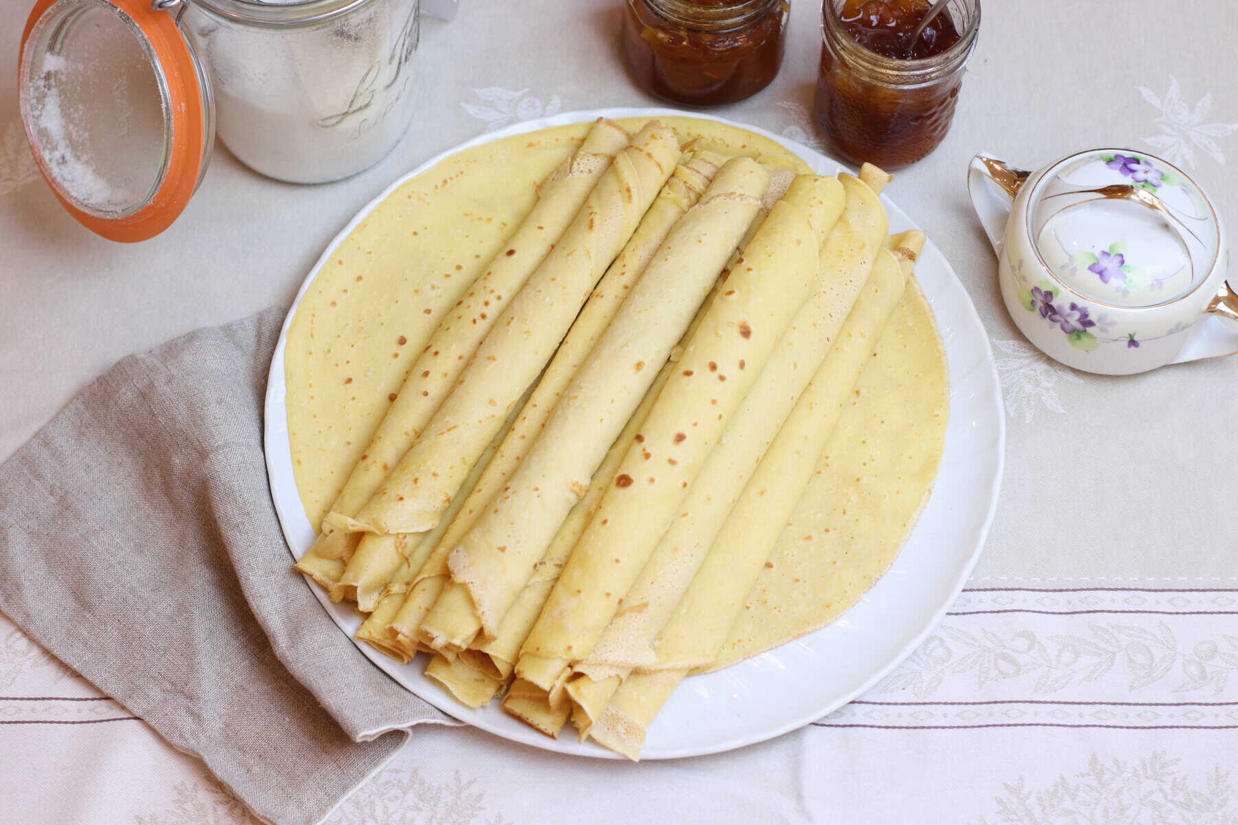 Fluffy French crepes topped with powdered sugar on a white plate, accompanied by jam jars, a vintage teapot, and a linen napkin for a cozy French breakfast or brunch.