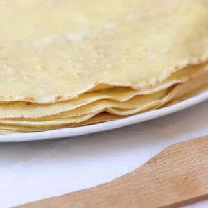 Thin French crepes stacked on a white plate with a wooden spatula nearby, showcasing classic French gouter or dessert options.