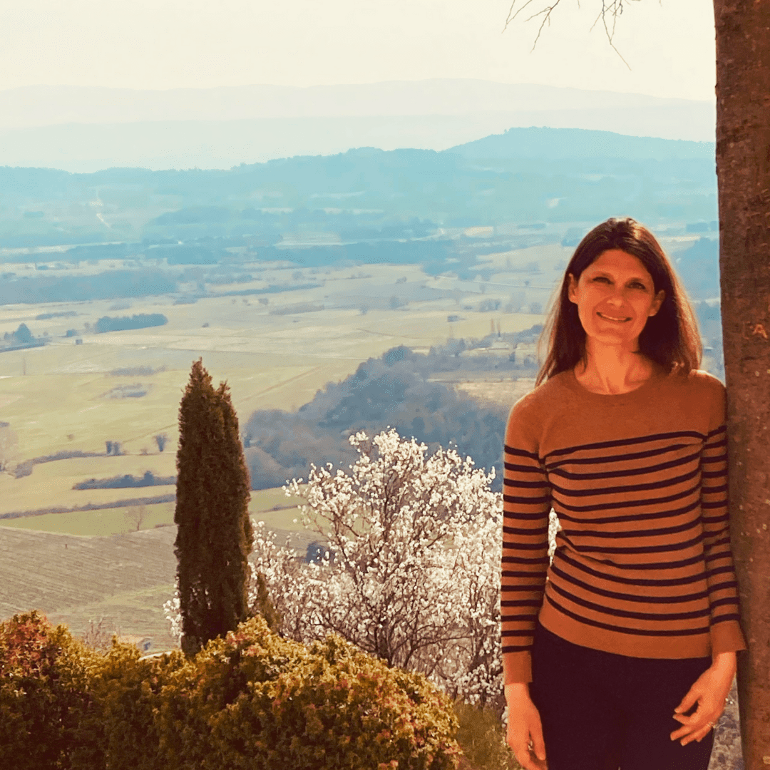A woman standing outdoors with a scenic French countryside view featuring rolling hills and blooming spring trees in the background.