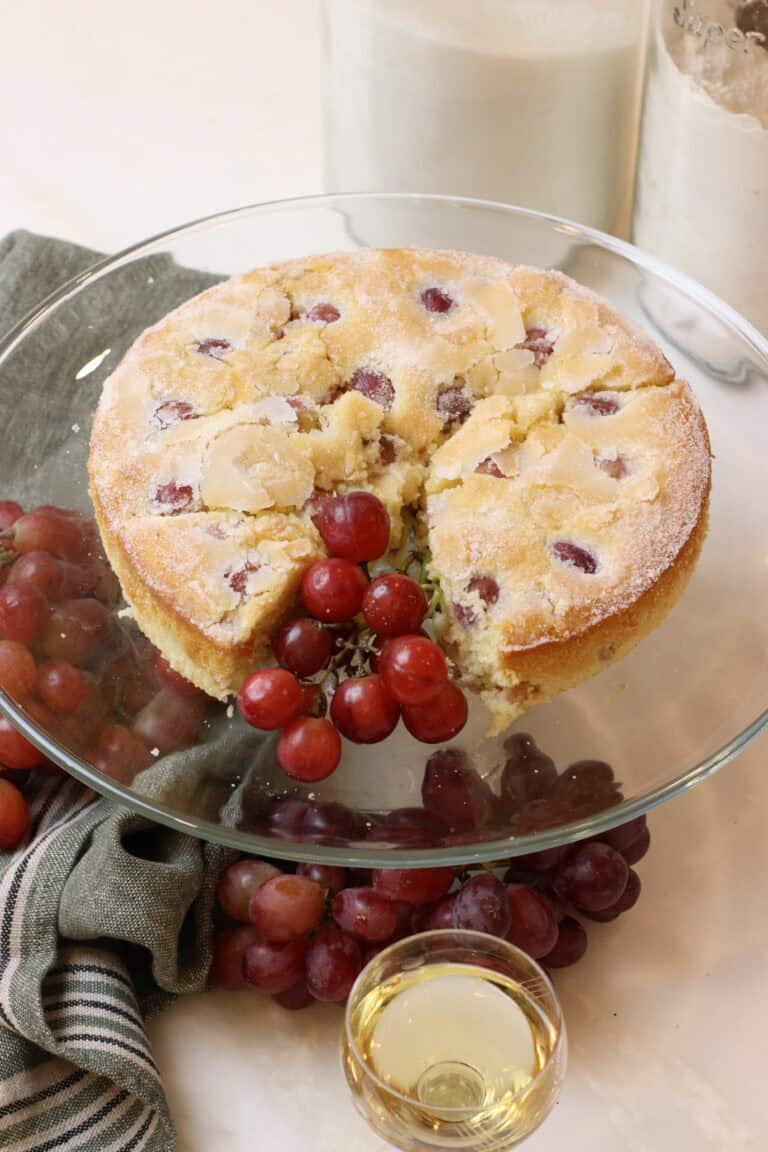 Freshly baked french wine cake with sugar, served on a glass cake stand with red grapes and a glass of white wine, embodying French dessert elegance.