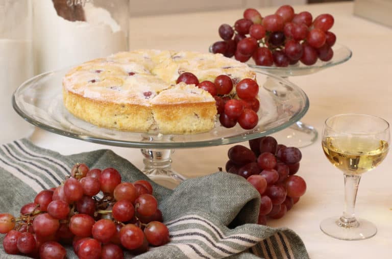 Grape cake on a cake stand surrounded by grapes and a glass of wine