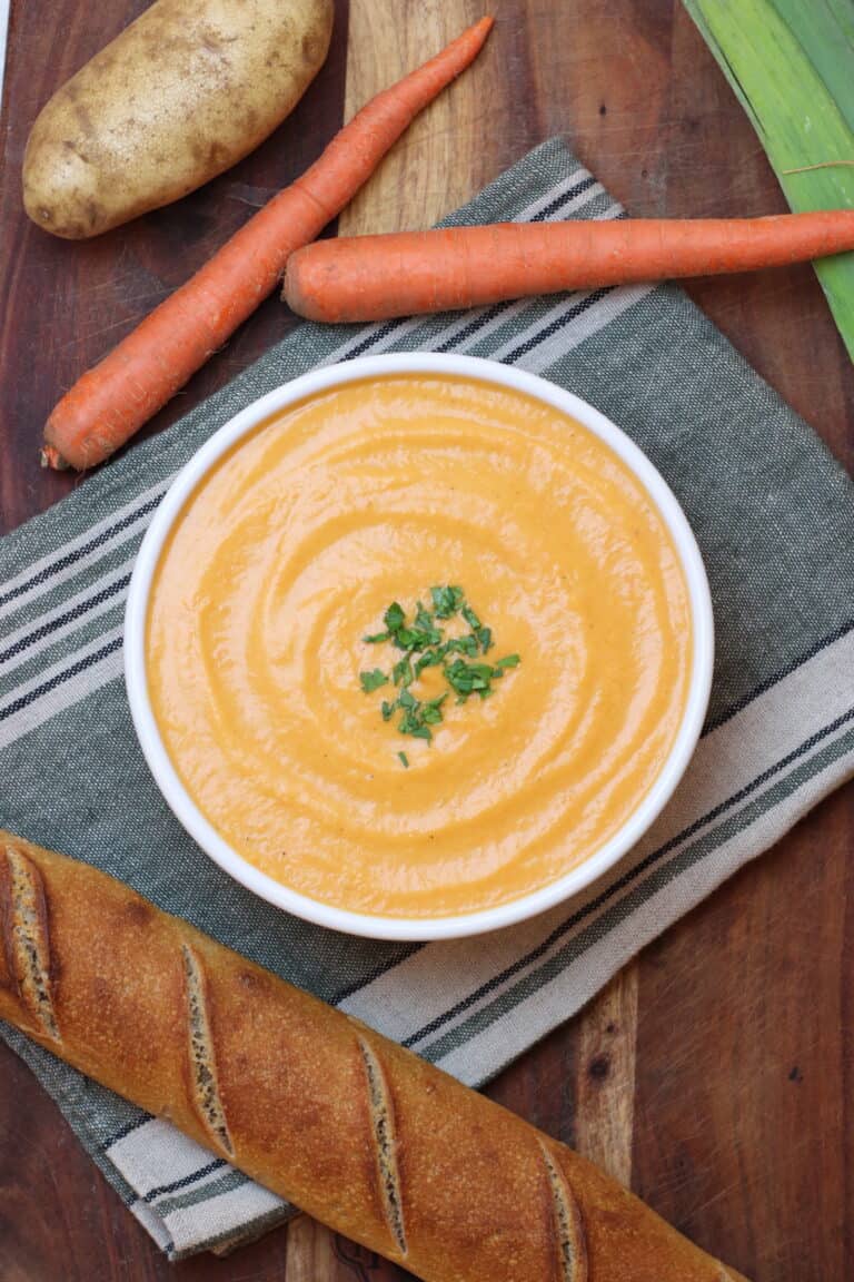 Freshly made vegetable potage soup served in a white bowl garnished with herbs, surrounded by carrots, a baguette, and potatoes for authentic French cuisine.