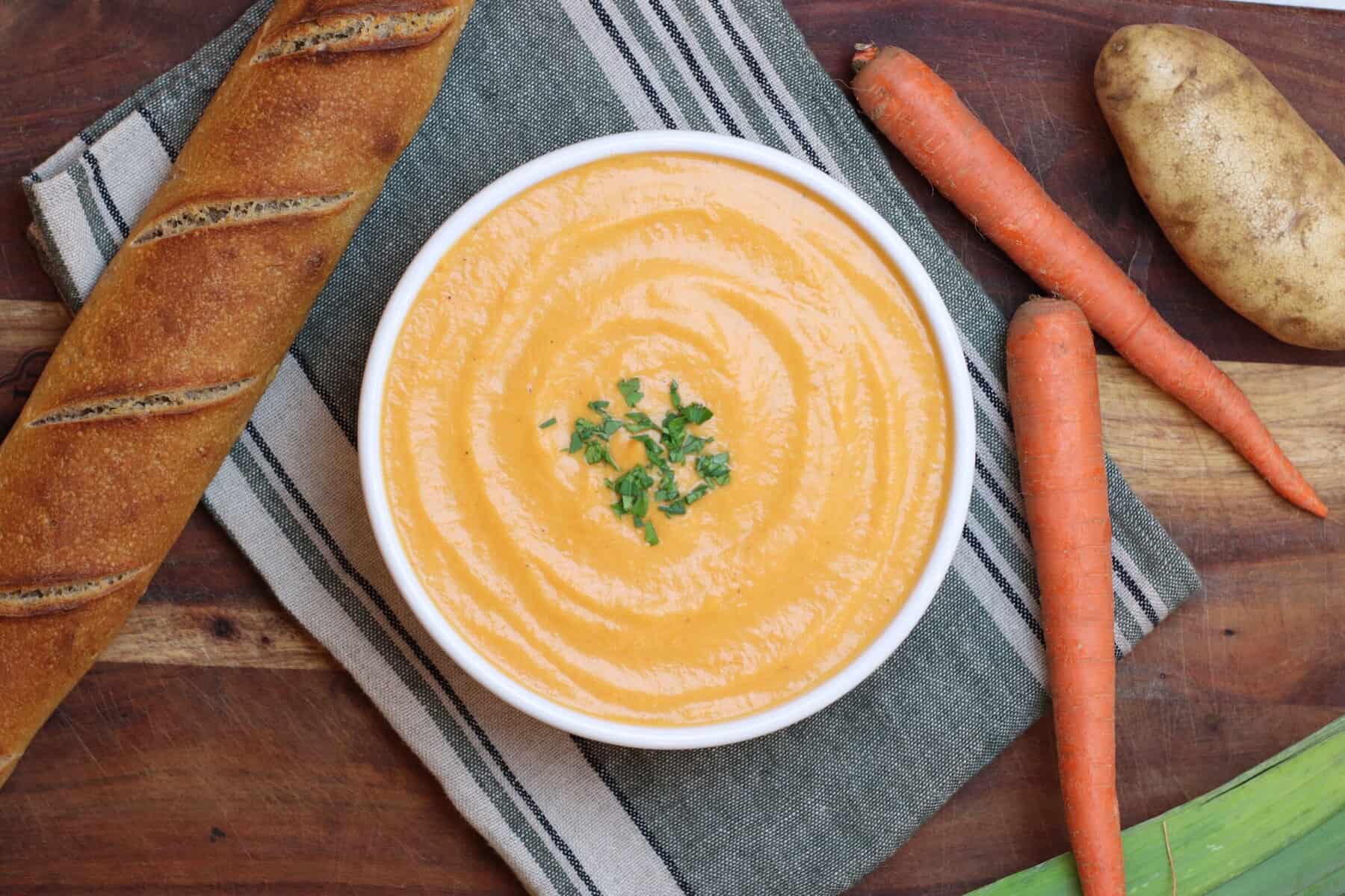 Overhead view of a bowl of dark yellow orange potage soup. Leeks carrots and potatoes along with a baguette are adjacent.