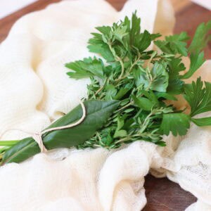 Bundle of herbs tied with a string on cheesecloth on cutting board.