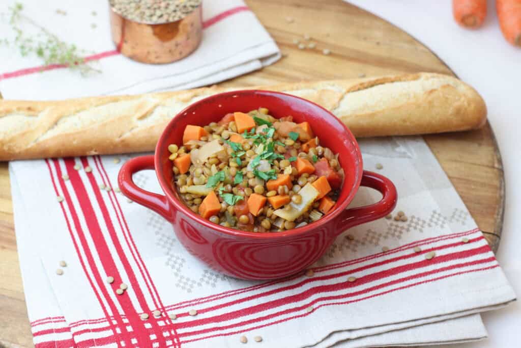 Hearty vegetable lentil soup in a red bowl with chopped herbs, served alongside a baguette on a wooden board, perfect for a cozy French-inspired meal.