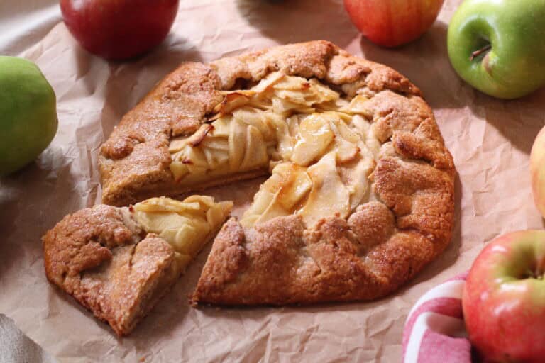 Freshly baked apple galette with a rustic crust, surrounded by colorful apples on a parchment paper surface. Perfect for French-inspired desserts and cozy gatherings.
