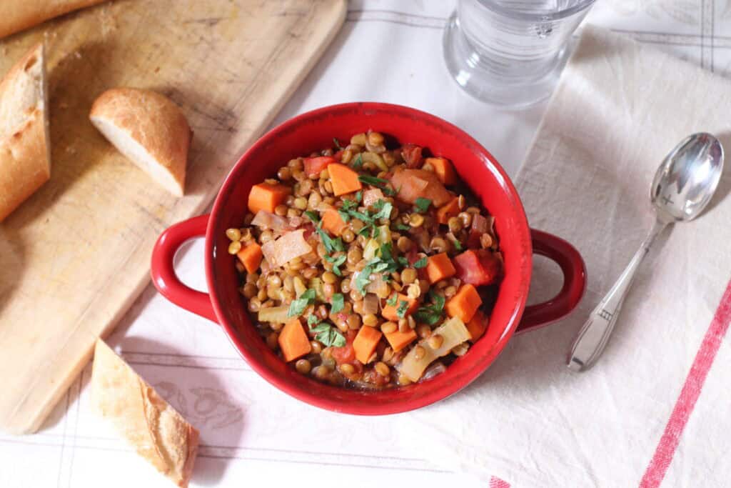 Hearty lentil vegetable soup served in a red bowl on a rustic table with bread and glass of water, showcasing cozy French-inspired home cooking and comfort food.