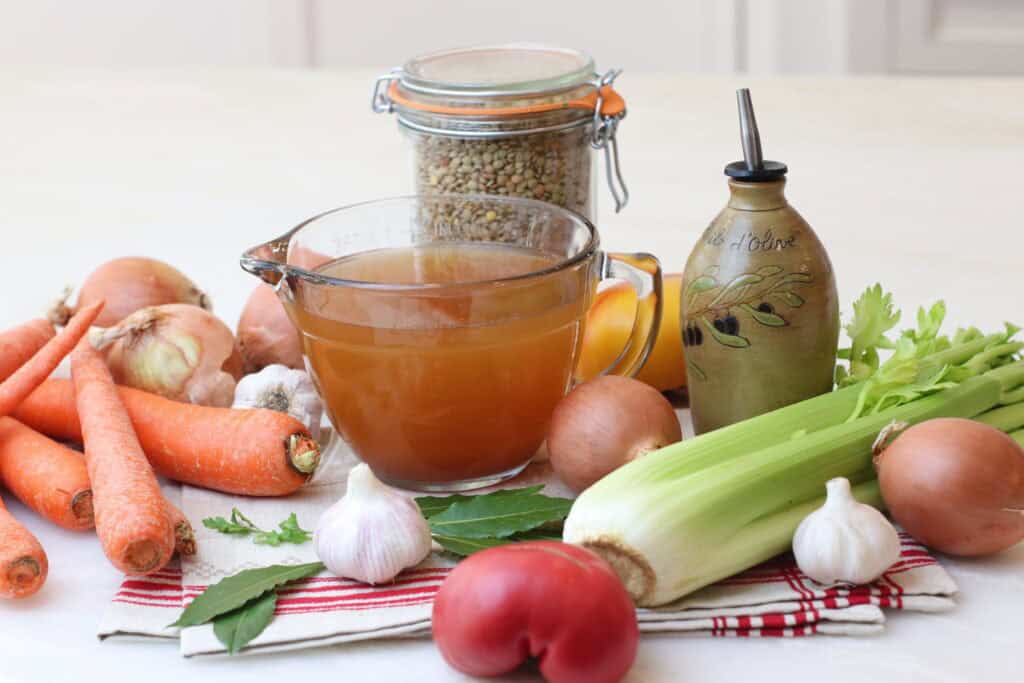 Fresh homemade vegetable broth with carrots, garlic, onions, celery, and herbs on a white kitchen table, capturing rustic French cooking elements.