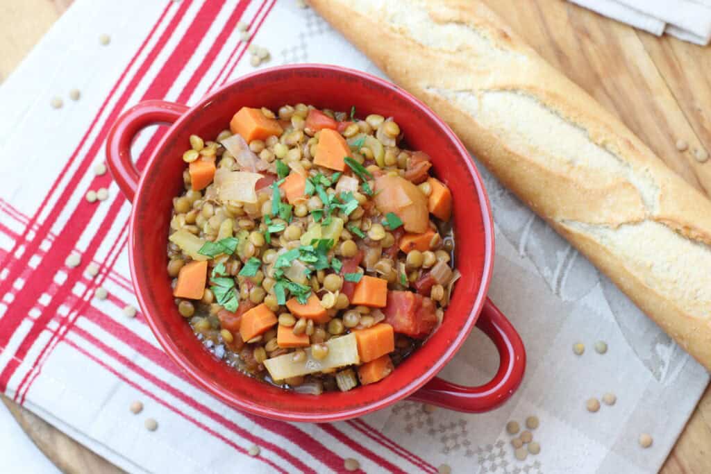Hearty vegetable lentil soup in a red bowl with chopped parsley, served with a baguette, on a rustic wooden table with a red and white checkered cloth, perfect for a cozy French-inspired meal.
