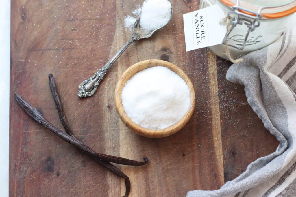 Vanilla bean pods, a silver spoon with vanilla powder, a wooden bowl filled with white sugar, and a jar labeled "Sucre Vanille" on rustic wooden surface.