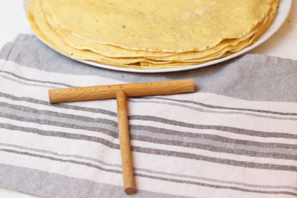 Delicious homemade crepes stacked on a white plate with a striped cloth and a small wooden cross, capturing the essence of French culinary traditions and cozy kitchen aesthetics.