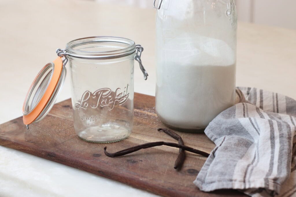 Homemade vanilla bean extract in a mason jar and flour in a glass jar, set on rustic wooden board with vanilla beans, grey striped cloth, and flour, showcasing classic French-inspired baking ingredients.