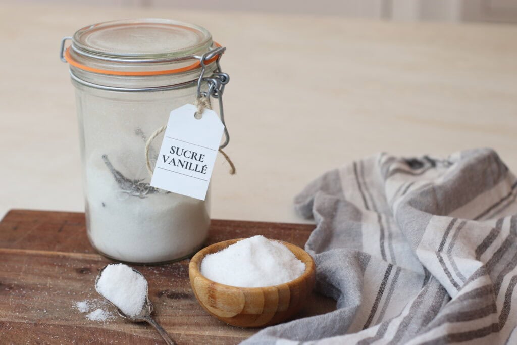 Sweet vanilla sugar in a glass jar with a vintage clasp, a small wooden bowl of granulated sugar, and a striped linen cloth on a rustic wooden surface. Perfect for baking or traditional French kitchen decor.