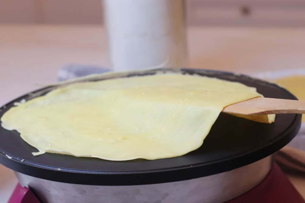 Thin crepe being cooked on a black electric skillet with a wooden spatula, showcasing classic French cuisine and homemade cooking.