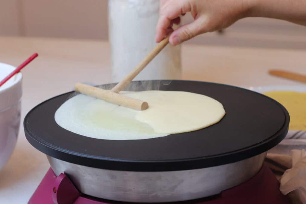 Light-colored crepe batter being spread on a non-stick electric crepe maker using a wooden spreader.