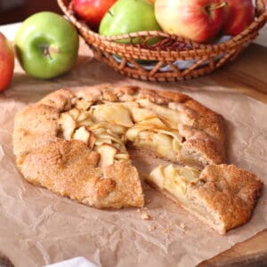 Fresh homemade apple galette on parchment paper with a basket of colorful apples in the background, showcasing rustic French pastry and seasonal fruit.
