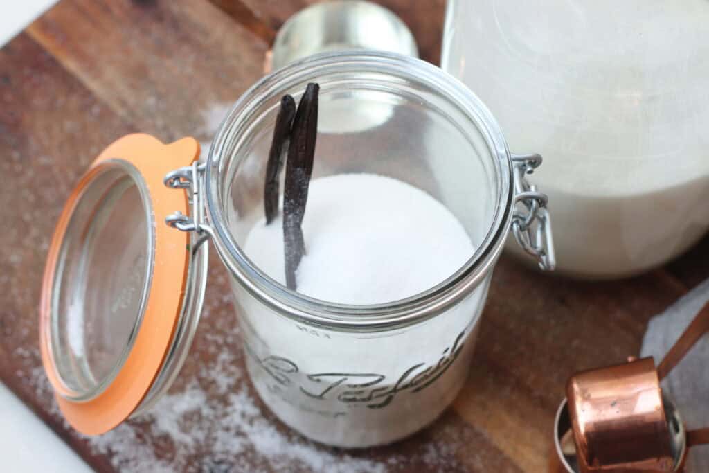 Cream of tartar in glass jar with black tongs, sugar, and baking supplies on rustic wooden surface, ideal for French baking and cooking.