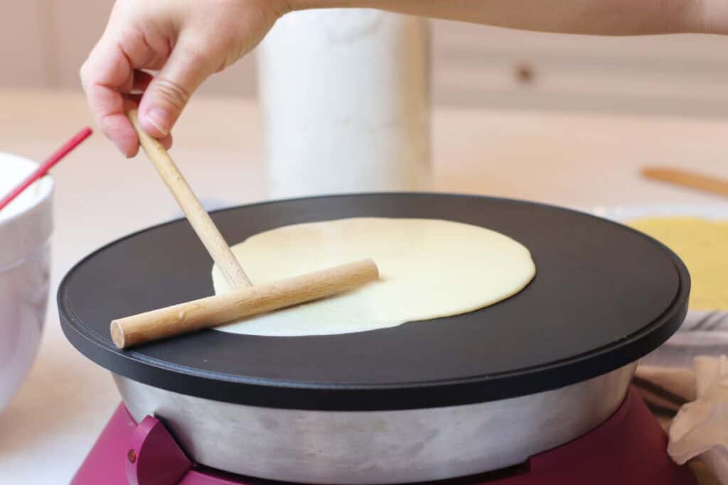 Crepe batter being spread on a non-stick crepe maker with a rolling pin, showcasing French culinary tradition and home cooking.
