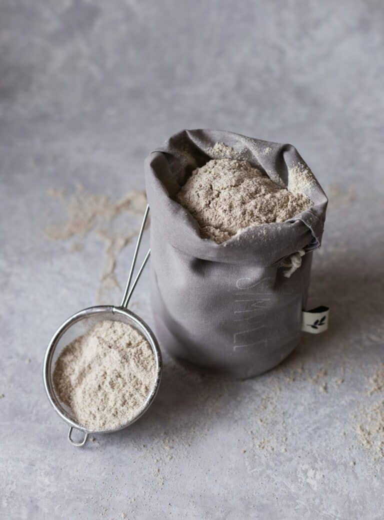 Fine flour in a gray fabric bag with a metal sifter, on a textured gray surface, ideal for French baking and pastry making, rustic baking ingredients, Quaint French Living.